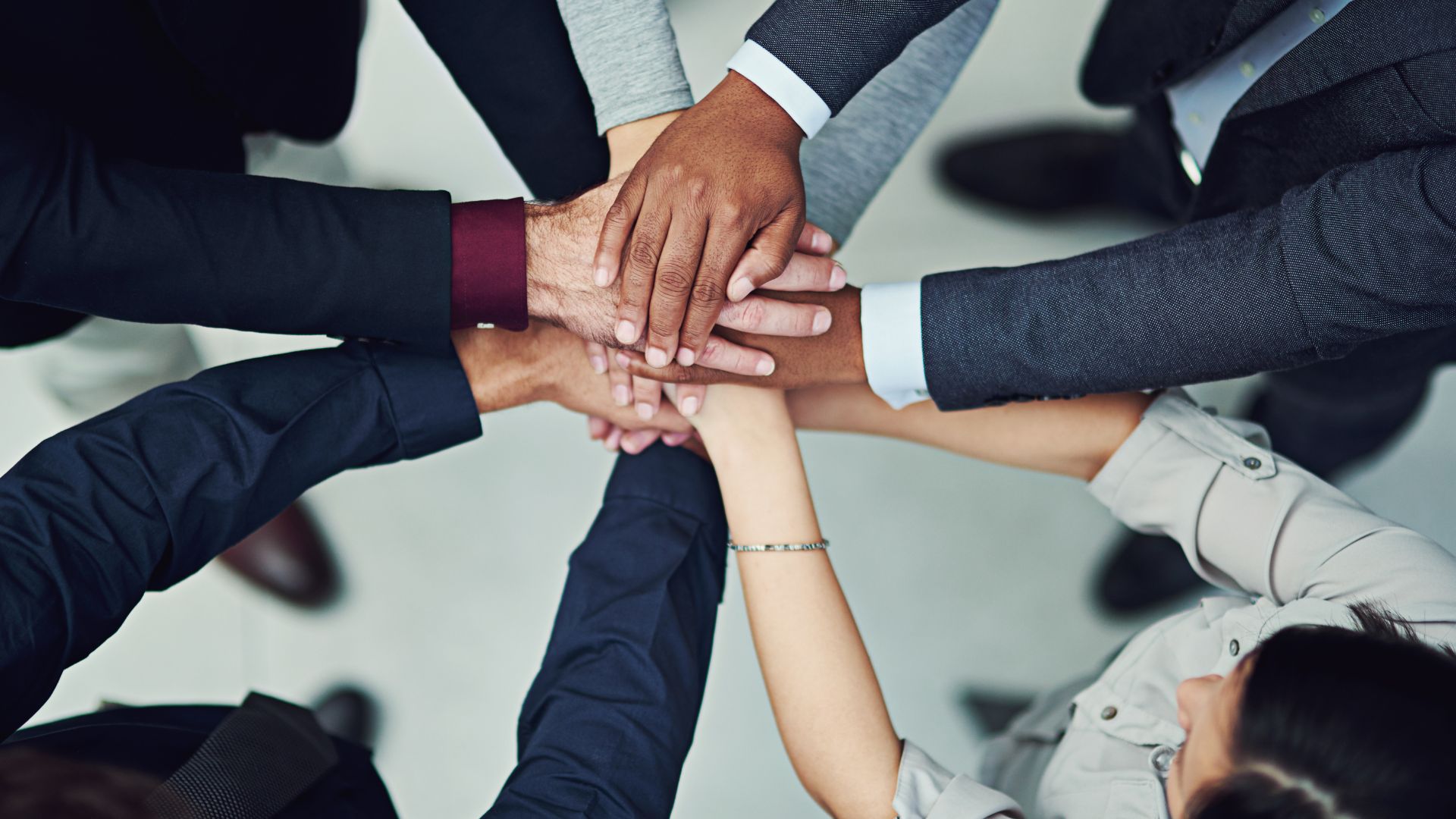 A group of professionals standing in a circle, stacking their hands together in a display of teamwork and unity. The individuals are dressed in business attire, symbolizing collaboration and partnership